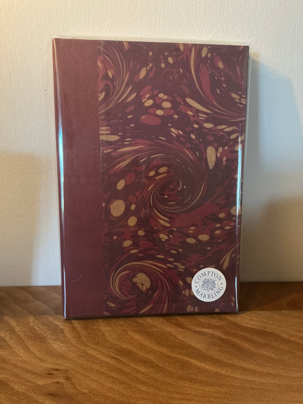 An A5 hardback journal standing on a wooden shelf, with a deep red cloth spine and a hand marbled cover of red and gold swirls.