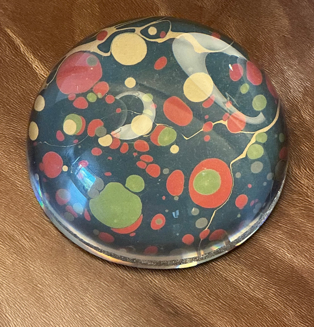 A domed glass paperweight on a wooden table, over hand marbled paper in dark teal with large red, yellow and green spots.