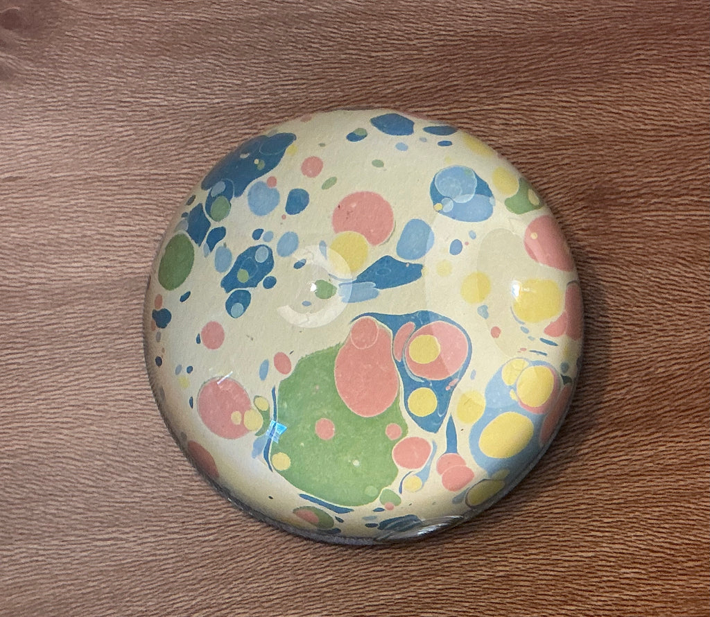 A domed glass paperweight on a wooden table, over hand marbled paper in cream spotted with blue, red and green.