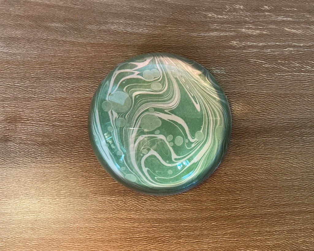A domed glass paperweight on a wooden table, over hand marbled paper in green with pale swirls running through it.