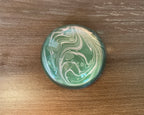 A domed glass paperweight on a wooden table, over hand marbled paper in green with pale swirls running through it.