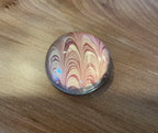 A domed glass paperweight on a wooden table, over hand marbled paper in cream with feathered arcs of deep red.